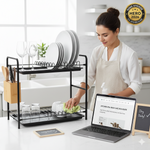 A 2026 home kitchen scene where a woman is cleaning a black metal dish drainer rack with a spray bottle and cloth, next to a laptop displaying a cleaning guide.